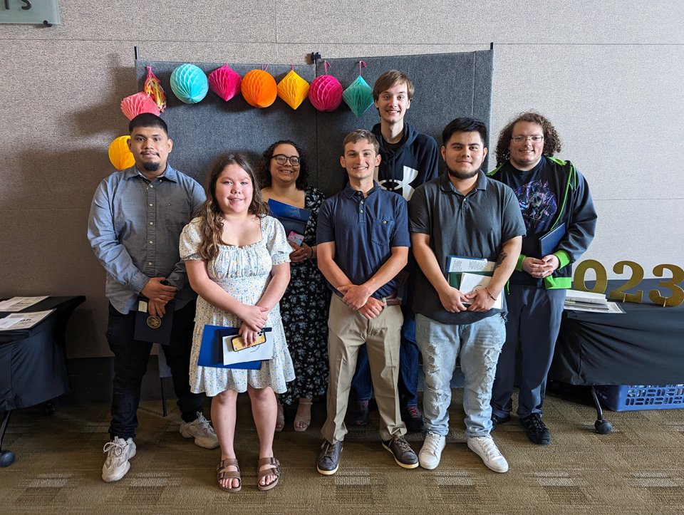 seven adult students standing against backdrop holding diplomas